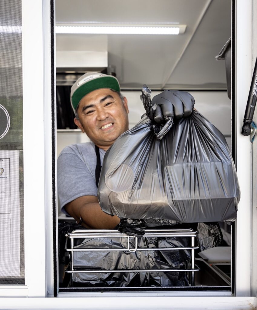 A man wearing a hat lifts a large black trash bag while standing in the back of a food truck. He is smiling slightly and facing the camera.