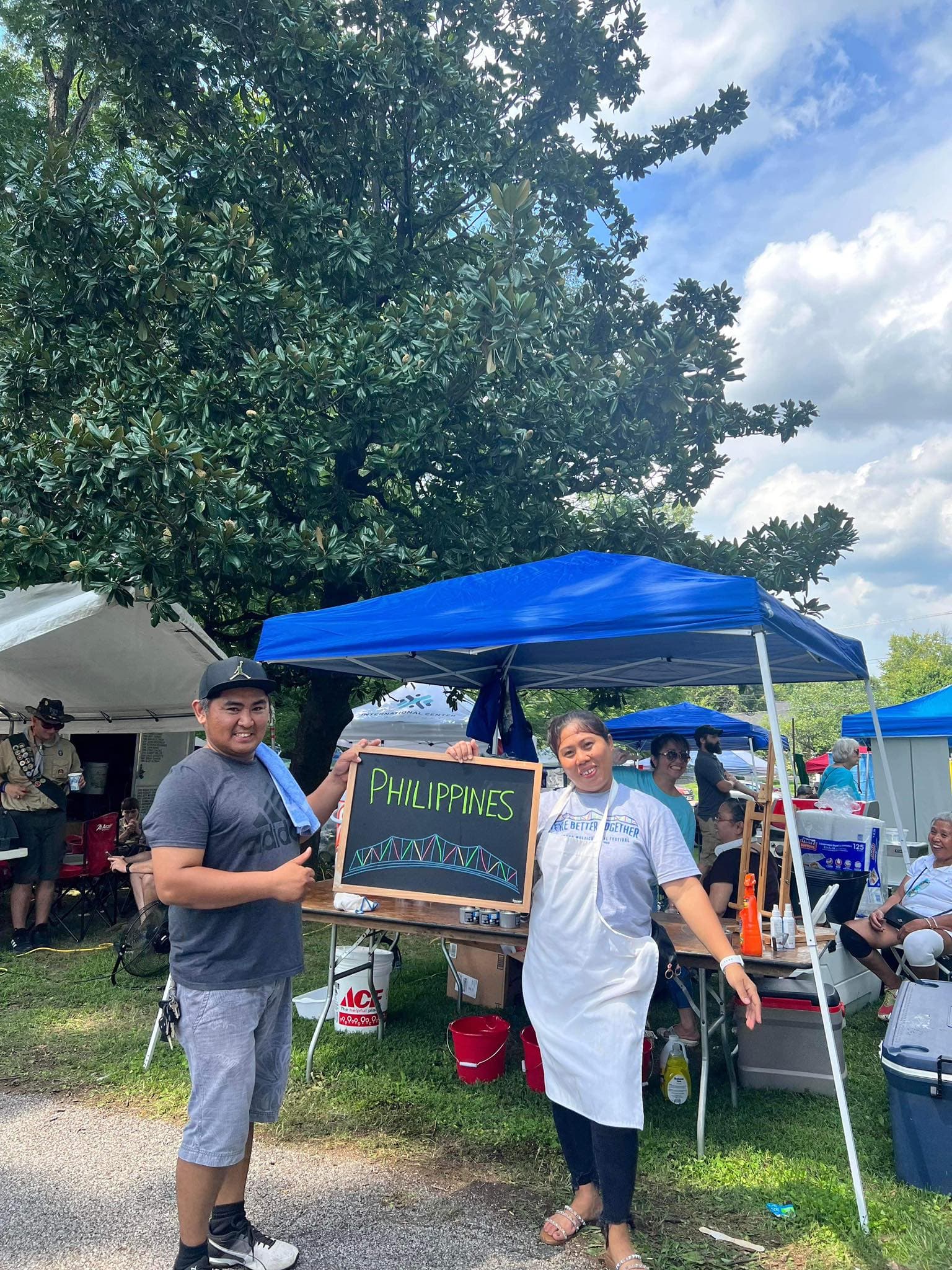 Two people standing under a blue canopy tent at an outdoor market. One is holding a chalkboard sign that says “Pancit & Lumpia.” Tables, tents, and a tree are visible in the background.